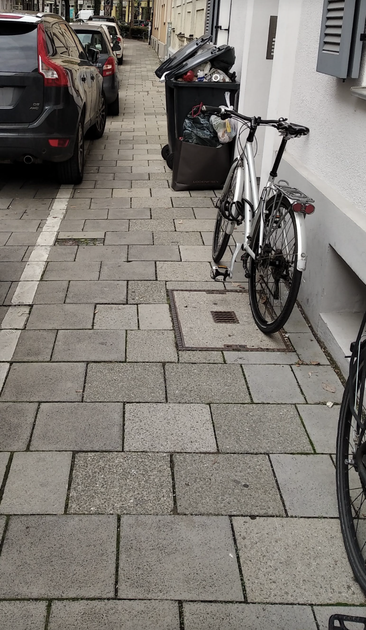 Münchner Gehweg (mit klassischer Münchner Gehwegplatte).

Links parken Autos drauf (anscheinend legal, es gibt eine Begrenzungslinie, die aber von den Parkern überschritten wird). Rechts an der Hauswand stehen ungelehrte, halb aufgeklappte Mülltonnen, weiter vorne ein Fahrrad.

Zwischen Auto und Mülltonne sind ca 2 Münchner Gehwegplatten (a 35cm) Platz, also etwa 70cm.