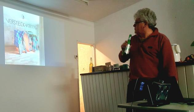 Ein Innenraumfoto zeigt eine Person, die vor einer weißen Wand steht, auf der ein Bild projiziert wird. Die Person trägt ein rotes Hemd und eine dunkle Hose und hält eine grüne Flasche in der rechten Hand. Das projizierte Bild zeigt eine farbige Darstellung von Objekten. Im Vordergrund befindet sich ein Schrank mit einer Tischplatte, auf der verschiedene Gegenstände wie Flaschen und Behälter stehen. Ein Projektor ist auf einem Tisch vor dem Schrank platziert. Auf der projizierten Folie steht de…