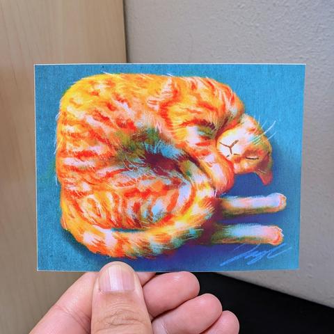 Photo of a left hand holding a rectangular sticker. The sticker is a matte painting of an orange cat shaped like a shrimp.