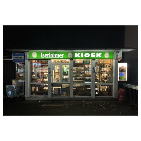 Ein kleiner Kiosk mit Flachdach in der Nacht. Frontal fotografiert. Die Gesamte Front ist aus Glas. Verkaufs-Artikel stapeln sich im Fenster. Unter dem Dach hängt über die ganze Breite des Gebäudes ein grünes leuchtendes Schild. Mit weißer Schrift steht darauf „Iserlohner Kiosk“ 
