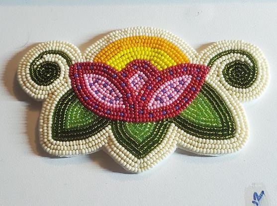 a beaded hairpiece made by kiamichi for her sister. on an ivory colored background, sits a 3-petaled flower in pink, purple, and red opening beneath the sun in yellow and gold. under the flower are three shiny vibrant green leaves with rolled-up vines extending to the right and left of the flower in the center. 