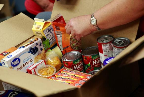 A hand packs assorted shelf-stable foods — canned pasta sauce, canned fruit, cereal bars, rice, and boxed sides — into a large cardboard food-donation box.