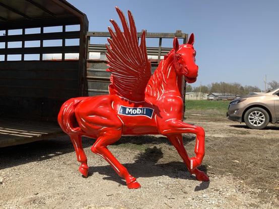 A three-dimensional horse sculpture that is almost life-size is painted red to look like the Mobile one Pegasus. It is near a horse trailer. side view. 