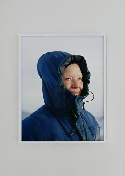 Ingelijste foto van Catherine Lemblé. Een vrouw met felblauwe jas en met kap op kijkt voor zich uit, ze heeft felblauwe ogen en glimlacht. Ze staat voor een witte achtergrond die een sneeuwlandschap suggereert. 