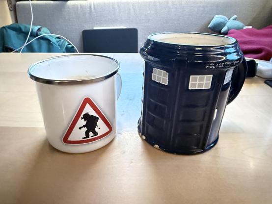 Zwei Tassen stehen auf einer Holzoberfläche. Die linke Tasse ist weiß und zylindrisch. Auf der Tasse befindet sich ein gelbes Warnschild mit einem schwarzen Symbol eines laufenden Trolls. Die rechte Tasse ist dunkelblau und hat die Form eines Hauses mit einem Henkel. Es gibt Text auf der Tasse, der „PUBLIC POLICEBOX“ lautet. Im Hintergrund ist ein Teil eines Sessels und ein roter Stoff zu sehen.