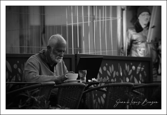 A quiet, momentary portrait in black and white: an older man with a short white beard sits alone at an outdoor cafe table, leaning slightly toward a laptop while holding a small coffee cup. The light sculpts his face and bald crown, giving his skin and hair a soft highlight against midtone clothing; the laptop’s dark screen makes his absorbed posture more pronounced. The foreground wicker chair and the patterned cafe partition — a row of stylized leaf cutouts — frame him horizontally, while a band of vertical metal bars and window reflections behind him add rhythm and depth.
Selective focus keeps the man and his immediate table crisp while the background dissolves into soft blur; a blurred standing figure or mannequin in a coat watches from the back, adding a faint, uncanny echo of presence without distracting. Small details — the saucer, a sugar packet or napkin, the slightly turned wrist — convey ritual and concentration: a familiar public solitude, half work and half repose.
In monochrome the scene reads as both timeless and contemporary: the classical stillness of coffee and contemplation set against the modern object of the laptop suggests a quiet negotiation between tradition and technology. Compositionally, the image uses leading lines, contrast, and shallow depth of field to isolate a private act within a public space, evoking themes of urban solitude, routine, and the gentle dignity of ordinary life. 

