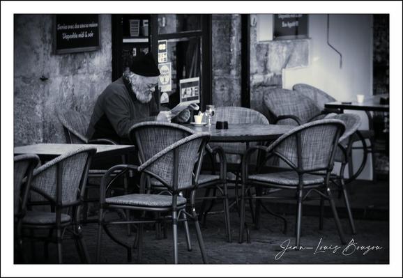 The image, presented in black and white, depicts a scene at an outdoor cafe. In the foreground are several tables and chairs. The chairs are made of woven material with dark frames. The tables are set with minimal items like salt and pepper shakers, and one table has a small cup and glass.
The central subject is an older man sitting at one of the tables. He is wearing a dark hat and a dark jacket. He has a full white beard and appears to be deeply engrossed in reading a newspaper. His posture suggests concentration and solitude.
Behind him, the cafe's building provides a backdrop. A sign hangs on the wall offering "Services avec des sauces maison" (Service with homemade sauces), indicating it is a food establishment. The building's texture appears to be stone.
The image has a quiet and slightly melancholic atmosphere. The empty tables and the solitary figure create a sense of stillness. The monochrome palette further reinforces this feeling of calm and introspection. 

