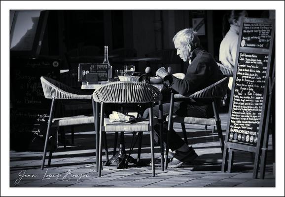 A quiet, high-contrast black-and-white photograph: an elderly figure sits alone at a small round café table, bathed in a shaft of bright light that turns their hair into a halo and carves long, crisp shadows across the paving. The scene is intimate and textural — the woven organic pattern of the café chairs, the ridged ceramic cup, a half-full bottle and glass catching specular highlights, and a stack of napkins or papers tucked under the table. To the right, a tall chalkboard menu with handwritten lines provides a vertical anchor and a hint of place without spelling it out.
The composition is driven by geometry and light. The circular table and chairs form a soft center against the hard, linear tiles and the rectangular menu board; strong sidelight sculpts the subject’s silhouette and separates foreground detail from a darker background. Small gestures — hands close together at the table, shoulders slightly curved — convey concentration and ritual, as if the person is engaged in a slow, deliberate task: stirring, reading, or simply holding a warm cup.
Culturally and symbolically, the photograph reads like a quiet study in solitude and time. TRendered in monochrome, the image emphasizes contrasts: light and shadow, texture and smoothness, presence and emptiness — suggesting themes of memory, endurance, and the paused rhythm of a single afternoon. 