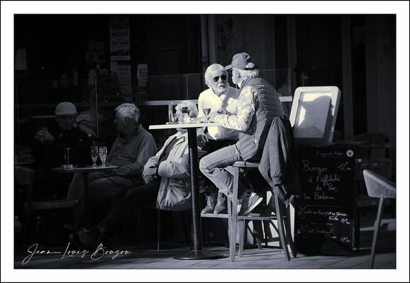 
A quiet, cinematic street café frozen in monochrome. Two men sit high on bar stools at a small round table center stage; one leans forward, the other turns slightly toward him — their conversation looks intimate and absorbed. The nearer figure wears sunglasses that catch a bright highlight, a light collar against a darker jacket. On the tabletop are the small rituals of café life: a cup, a bottle, ashtray or small bowl, the casual clutter that signals a pause in the day.
The photographer frames them with strong side light: faces and hands are picked out in sharp relief while the background slips into shadow. Behind and around them, other patrons are dimmer shapes—an older woman seated lower, a chalkboard menu leaning against the wall — which grounds the scene in a public but private moment. Textures are tactile: the grain of the pavement, the smooth shine of glass, the soft folds of coats come through more insistently because the image is black and white.
Symbolically it reads as a study of everyday intimacy and urban ritual — a small island of connection amid the anonymity of street life. Artistically, the picture uses high contrast and selective lighting to create depth and focus, turning a common café encounter into a quiet tableau about conversation, observation, and the small performances of social life.
