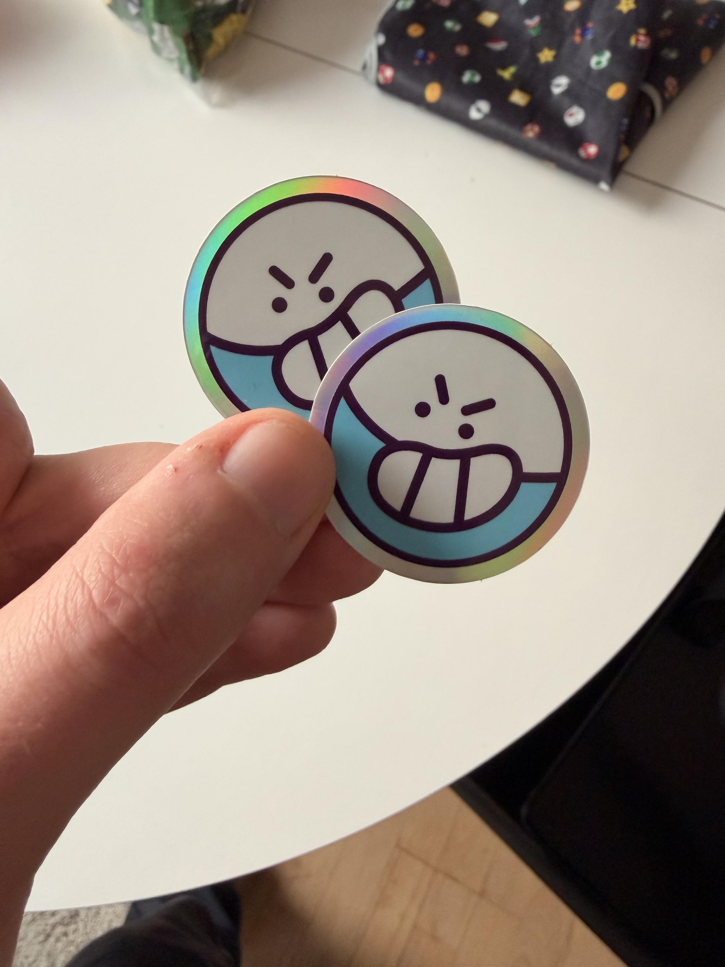 A hand holding two robb stickers with iridescent backdrops. They show a drawing of a shiny electrode with a wide grin.