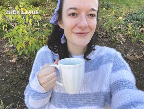An outdoor selfie by Lucy LaRue wearing a striped light blue and white fuzzy sweater and a blue hankie in her brown curly hair, holding a white coffee mug and smiling.