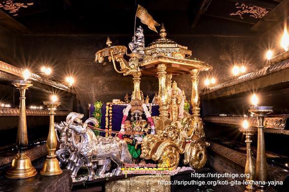 Wide shot of a temple altar with idol of Hindu deity Krishna dressed as Satyabhaamaa in green seated in the driver seat of a horse-drawn gilded chariot with a gold statue of Krishna (in standing pose) in the passenger seat. The chariot is drawn by four silver horses [screen left] and has an ornate roof with a small umbrella at its front. At the leading edge of the roof, behind the umbrella, sits Lord Hanuman holding up a gilded flag. A string of white and red banded flowers around Krishna (in the driver seat). Two rails of oil lamps on either side of the chariot, and two tall traditional oil lamps on both sides of the chariot in the front. Dark blue cloth background behind the chariot.