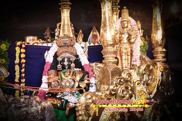 A temple altar with idol of Hindu deity Krishna dressed as Satyabhaamaa in green seated in the driver seat of a horse-drawn gilded chariot with a gold statue of Krishna (in standing pose) in the passenger seat. The chariot is drawn by four silver horses [screen left] and has an ornate roof with a small umbrella at its front. At the leading edge of the roof, behind the umbrella, sits Lord Hanuman holding up a gilded flag. The chariot is positioned on a gold-covered pedestal. A string of white and red banded flowers around Krishna (in the driver seat). A tall traditional oil lamps on screen left in front of the horses. A few other lamps in the background. Dark blue cloth background behind the chariot.