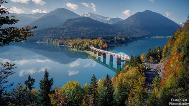 A breathtaking autumn landscape unfolds, showcasing a stunning lake nestled between rolling hills and majestic mountains. The lake, with its deep blue waters, mirrors the sky and surrounding scenery, creating a perfect reflection that enhances the serene atmosphere. The calm surface of the water adds a sense of tranquility and peace to the scene.

A long bridge stretches across the lake, connecting the two sides of the lush, forested hills. The bridge, with its elegant arches, adds a touch of h…