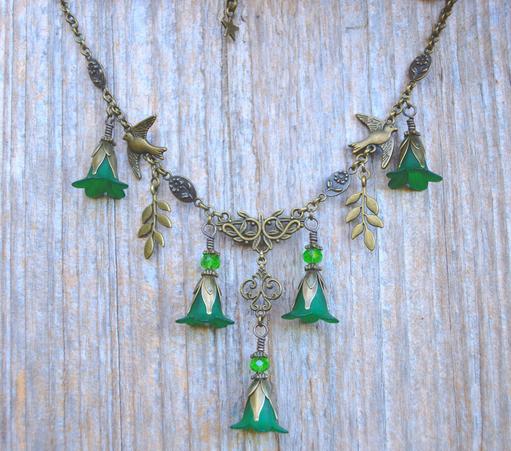 Close up of a bronze necklace with 5 dangling green flower pendants, litte birds in flight and willow leaves dangling from a v shaped chain. Background is light weathered wood.