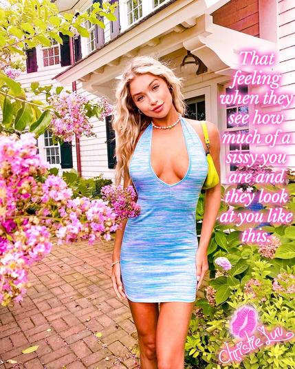 A woman in a flowing blue dress poses gracefully with a smirk on her face in front of a charming house, showcasing her vibrant style. "That feeling when they see how much of a sissy you are and they look at you like this."