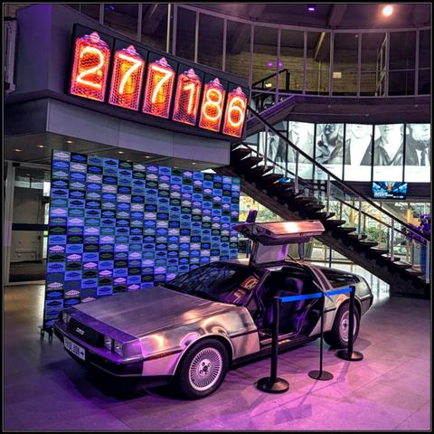 A DeLorean DMC-12. The type that is known for featuring in the Back To The Future movies. It is sitting inside the lobby of the Next Nature museum in Eindhoven, a blue retro patterned backdrop and a curved staircase behind it. There is an illuminated counter above it with the numbers  277186

The car is illuminated with pink and purple lights. The drivers side door is open and there is a ribbon in front of it to indicate we're not allowed to sit in it.