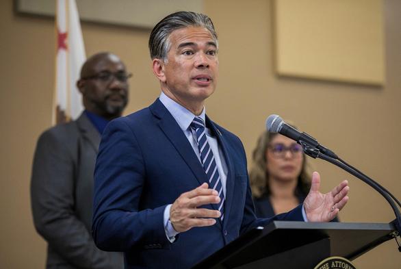 A man in a navy blue suit and striped tie speaks at a podium with a microphone, gesturing with his hands. Two people stand blurred in the background, including a man in a gray suit and a woman in glasses. A portion of a California state flag is visible behind them.
