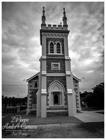 A black and white, low angle photograph of the historic Manaranga Church, featuring a prominent central tower with Gothic style pointed arch windows and battlements at the top, capped by two small spires.
The church facade is built from contrasting materials, possibly stone and render. The sky is heavily clouded, adding a dramatic, moody atmosphere.