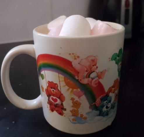 Care Bears mug, with bright CareBears and rainbows on a white background,  with white and pink marshmallows heaped on top of salted caramel drink in the mug. The mug sits on a black countertop.