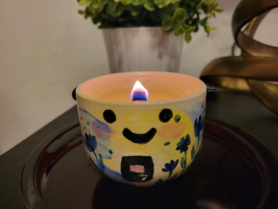Nighttime interior photo of a small lit candle. The candle wax is in a ceramic container that looks like a rice cooker, with two black handles and a square cook button/switch. It is hand painted in pastel colors, with a  smiling yellow face and blue flowers.