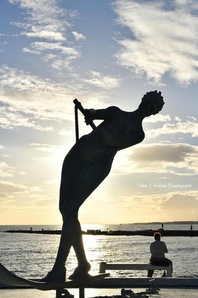 Imagen en formato vertical de la escultura 'La Venus' a contraluz. Cerca se ve a una persona sentada en un banco contemplando el atardecer y el mar. El cielo tiene cierta nubosidad.