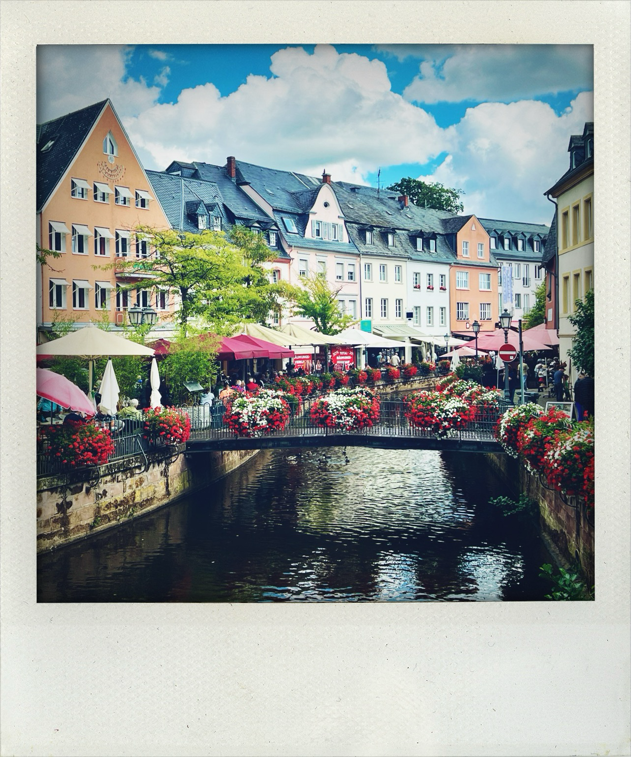 Foto zeigt eine Fußgängerbrücke über die Leuck, die mit Blumen geschmückt ist. Links Sonnenschirme , dahinter Altstadt. 