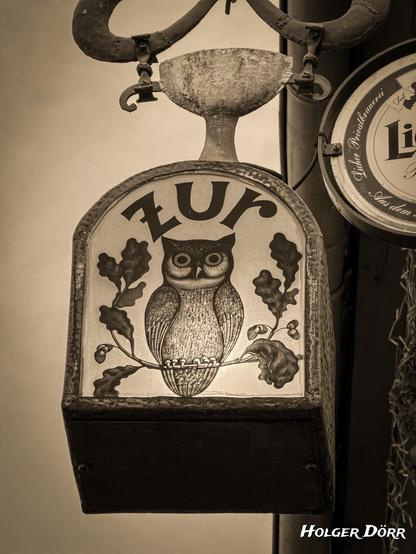 Sepiafarbenes Foto eines historischen Wirtshausschilds der Gaststätte „Zur Eule" in Laubach. Das beleuchtete Schild zeigt eine stilisierte Eule, umrahmt von Eichenblättern, getragen von einer alten, leicht verwitterten Metallhalterung. Die warme Tönung verleiht dem Motiv eine nostalgische, ruhige Atmosphäre und erinnert an die lange Geschichte des Gasthauses.