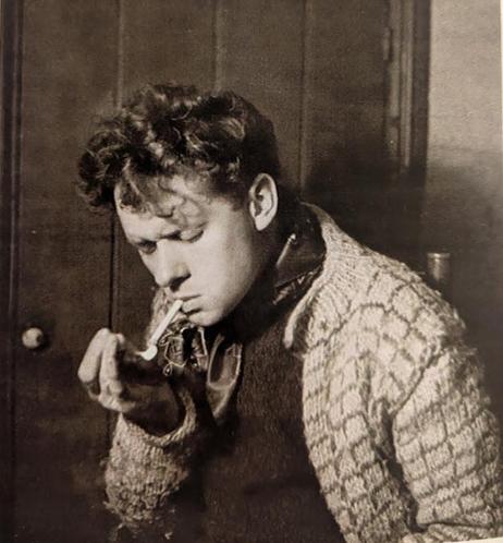 Nora Summer's iconic portrait of Dylan Thomas. Taken in Blashford in 1937, it was soon to gain an international circulation.

Dylanis is wearing a distinctive thick, cable-knit cardigan with a textured pattern, and what appears to be a scarf around his neck.

He was photographed from the side, looking downward with his eyes cast down, holding a cigarette to his lips. His hair is characteristically curly and somewhat disheveled.

https://en.wikipedia.org/wiki/Dylan_Thomas#/media/File:Dylan_Thomas_icon_Blashford_1937.jpg