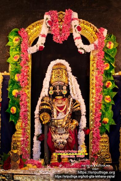 Idol of Hindu deity Krishna in red standing on a gold-covered pedestal, right hand in giving pose, left hand on front left waist; wearing gold shoes, gold palm coverings, gold crown, and lots of gold ornaments including large lotus-shaped medallion which hangs well below his waist. A string of white flowers around Krishna floor-floor. A gold arch around Krishna. A string of pink flowers hangs down along both sides of the arch, along with a string of green betel leaves (yellow flower on each leaf). A string of white flowers hooks from the left end at the top of the arch to the middle; likewise on the right end of the arch. Dark cloth filling the arch interior behind Krishna. Loose red roses and other flowers at Krishna's feet.