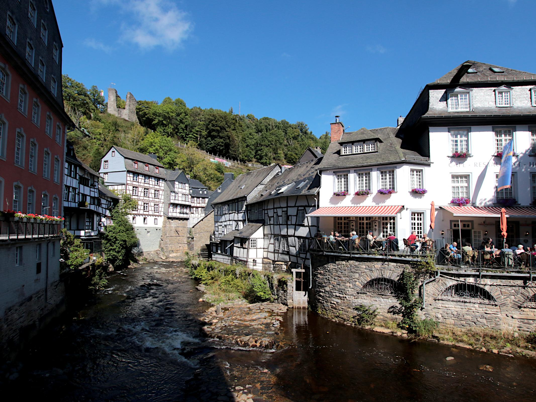 Das Bild zeigt einen Teil der historischen Altstadt von Monschau. In der Mitte des Bildes ist der Fluss Rur zu sehen, der an dieser Stelle von den Fundamenten der umstehenden Häusern eingefasst ist. Rechts neben einem Felsen im Flussbett ist  Schieber zu sehen, über den Wasser zu einem Haus abgeleitet werden kann. Dort ist ein Wasserrad einer alten Textilfabrik, das auf dem Bild aber nicht zu erkennen ist. Links im Bild kann man einen Teil des roten Hauses, einem Museum sehen. Rechts im Bild sieht man eine Terasse am Wasser, auf der Menschen in der Sonne sitzen. Im Hintergrund stehen noch einige Fachwerkhäuser und viel weiter hinten auf einer Anhöhe sieht man die Ruine einer alten Befestigungsanlage.