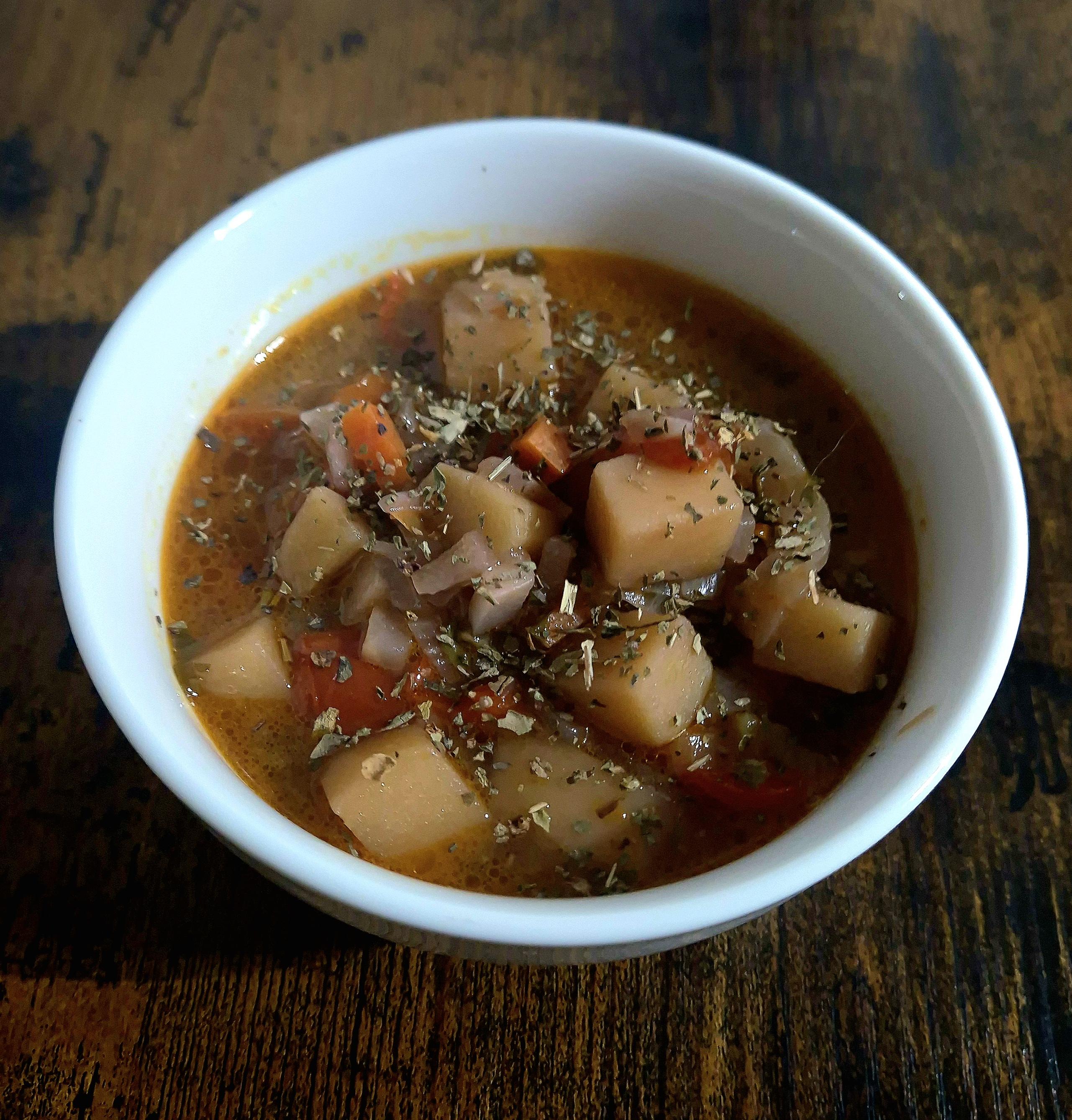 A white bowl filled with minestrone soup, showing diced potatoes, tomatoes, onions, and other vegetables in a reddish broth, topped with dried herbs. The bowl sits on a dark wooden surface.