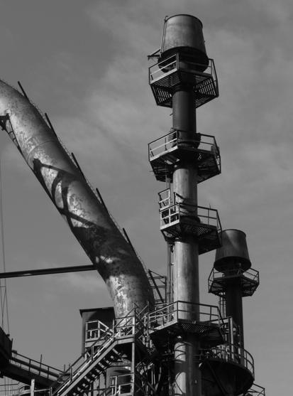 Part of the defunct Bethlehem Steel works, idle now for thirty years, that I think was part of the process used to clean the gases used to fuel one of the blast furnaces to which it is attached, but not part of the photograph. There is a large pipe on the left angled downward through which the gas flowed . To the right are two towers at the tops of which are large components that look like upside down buckets. Below those, are a number of hexagonal platforms spaced at intervals and supported by tall cylindrical poles. A stairway leads to the bottom platform closest to the foreground. Everything is made of metal. Sunlight and shadows fall upon the structure that stands starkly against the background sky. 