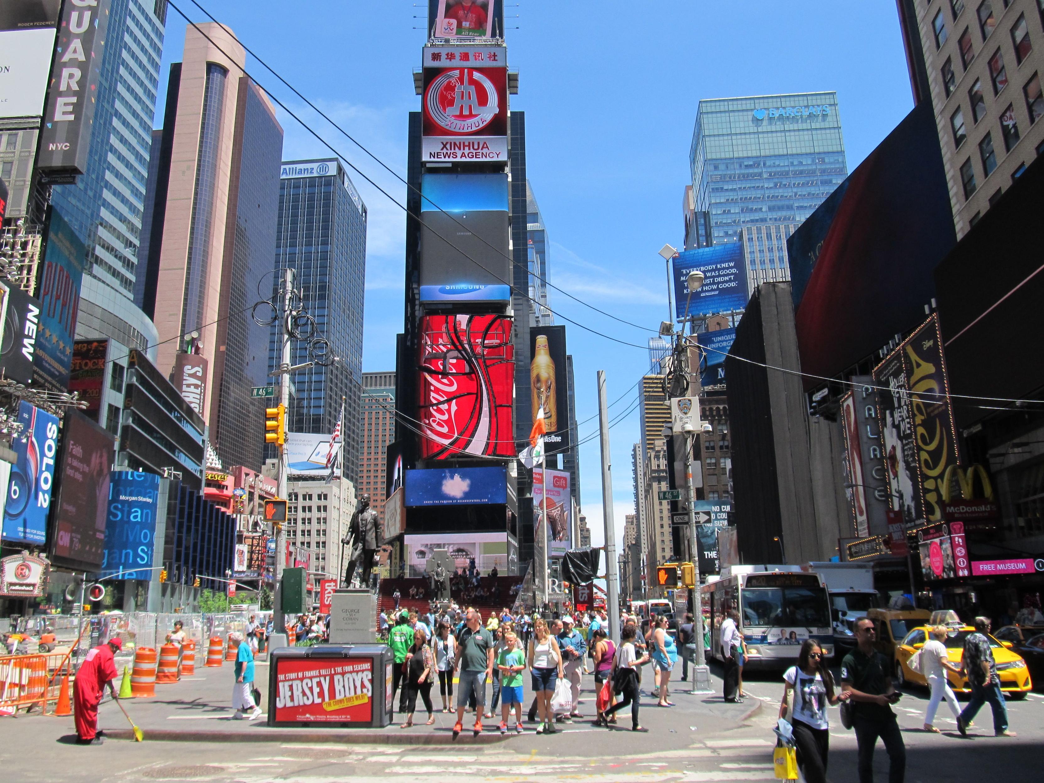 Eine Straßenszene, links und rechts moderne hohe Häuser, im Vordergrund spazieren Menschen, in der Mitte ein hohes, schmales Gebäude mit Leuchtreklame