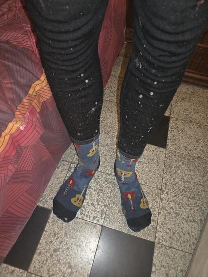 Deux pieds en chaussettes sont visibles sur un sol carrelé. Les chaussettes sont bleu foncé ornées d'illustrations de guitares. La personne porte égalementun legging noir qui est plein d’éclaboussures de boue. Le sol est recouvert de carreaux gris et noirs dans un motif de damier.