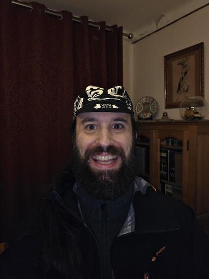 Un portrait rapproché d'une personne. La personne porte un casquette de cycliste noir avec un motif blanc. Elle a une barbe épaisse et foncée et sourit en montrant ses dents. Le fond est sombre et flou, avec des objets indistincts, dont un tableau et un meuble.