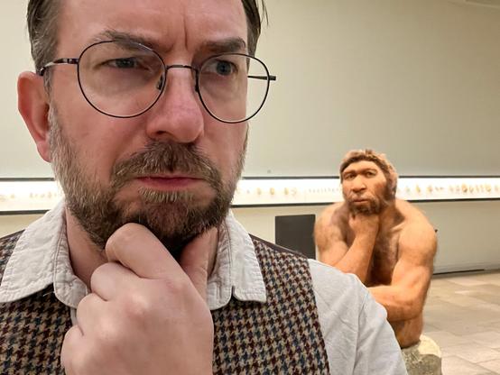 Selfie eines Mannes mit Bart, Brille und Weste, Daumen und Zeigefinger der linken Hand nachdenklich ans Kinn gelegt. Im Hintergrund die Rekonstruktion eines nackten, sitzenden Neanderthalers, den Kopf nachdenklich auf die rechte Faust gestützt.