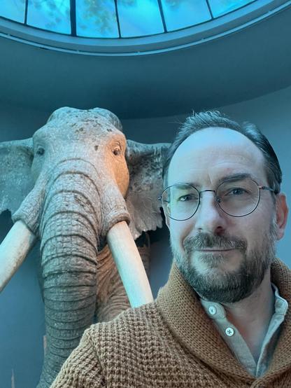 Selfie: Im Vordergrund ein Mann mit Bart, Brille und brauner Strickjacke, links im Hintergrund ein lebensgroß rekonstruierter Waldelefant, der frontal aus der Wand zu kommen scheint.