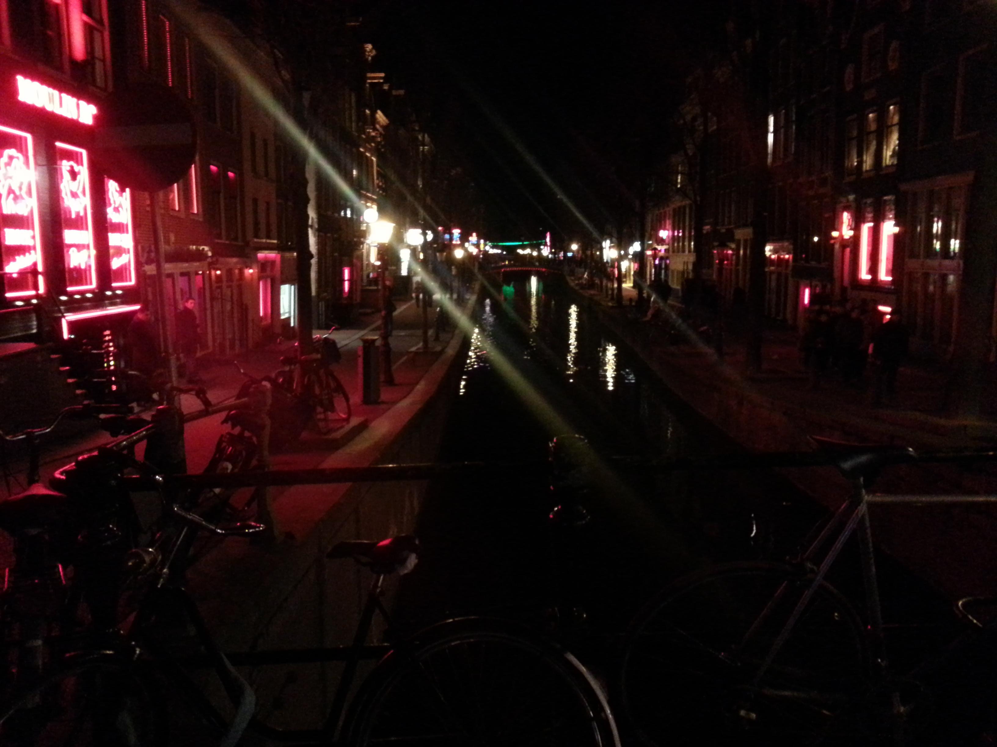 Bildbeschreibung von KI:
Das Foto zeigt eine nächtliche Szene an einem Kanal in Amsterdam. Die Umgebung ist dunkel, doch viele rote und pinke Lichter aus Schaufenstern und Leuchtreklamen erhellen die Strasse. Rechts und links des Kanals stehen alte schmale Häuser, typische Amsterdamer Bauweise. Das Wasser in der Mitte spiegelt die Lichter und wirkt ruhig. Auf der Brücke im Vordergrund stehen mehrere Fahrräder, die als dunkle Silhouetten zu erkennen sind. Auf beiden Seiten des Kanals gehen Menschen entlang, manche stehen und unterhalten sich. Die Stimmung ist lebhaft, aber gedämpft durch das nächtliche Licht. Die roten Leuchten verleihen der Szene einen warmen, leicht geheimnisvollen und touristischen Charakter.
