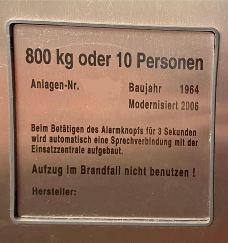 Metal plaque with information about an elevator, indicating a weight limit of 800 kg or 10 people. It provides details like the system number, year of installation (1964), modernization year (2006), and instructions for activating the alarm button to