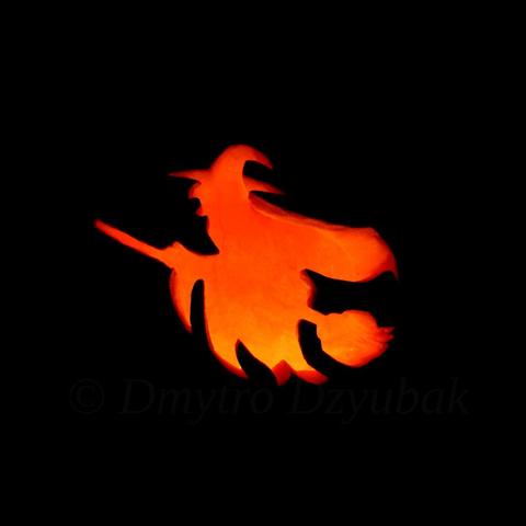 Witch wearing a black waving cloak and a pointed hat, flying on a broomstick is depicted on a carved pumpkin. Pumpkin with a candle inside at night. Lights off.