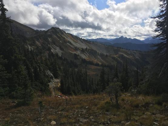 Ridge of a rugged mountain with more rugged mountains at the distance. Golden brown underbrush mixed with small groups of evergreen trees. The sky is cloud covered with occasional blue spots.