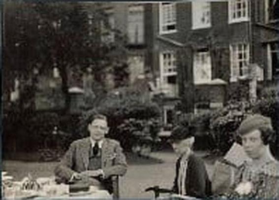 T.S. Eliot with his sister and his cousin

by Lady Ottoline Morrell
vintage snapshot print, 1934

The photograph is a vintage black-and-white snapshot from 1934, taken outdoors in what appears to be a garden or courtyard with tall brick buildings in the background. Three people sit at a table set for tea.

On the left, Eliot in a suit sits facing the camera, hands resting on the table. In the center, Eliot's cousin wearing a dark coat and hat sits slightly turned to the side, holding a cane. On the right, Eliot's sister in a patterned dress looks downward, her expression quiet and reserved.

https://en.wikipedia.org/wiki/T._S._Eliot#/media/File:Thomas_Stearns_Eliot_by_Lady_Ottoline_Morrell_(1934).jpg