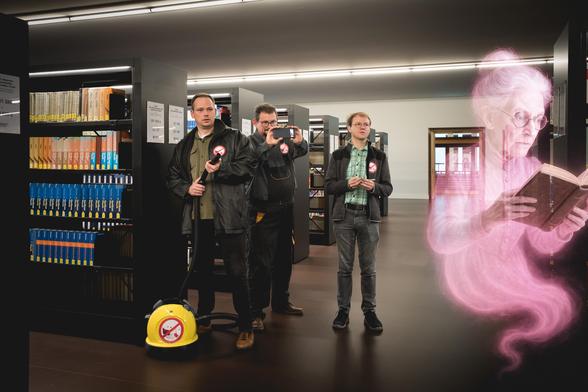 Ein Szene aus einem bekannten Filmklassiker, welcher in einer Bibliothek ist zu sehen. 3 Männer ausgerüstet mit allerlei Technik stehen im Freihandbereich der Bibliothek zwischen den Regal und starren recht überrascht, vielleicht sogar erschrocken auf eine Geistererscheinung einer Bibliothekarin.