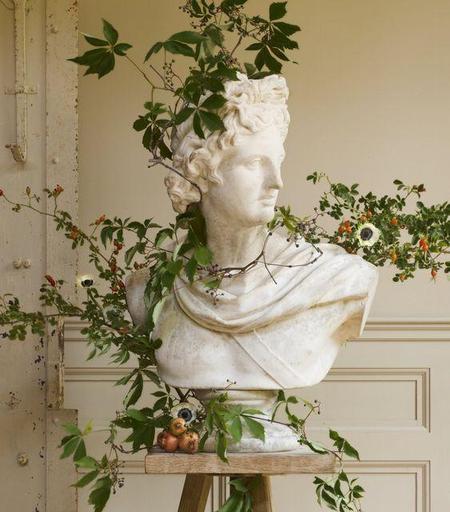 a bust of apollo, his head turned to the side looking to the right, , it is on a table, there are cut branches arranged around his handsome bust scukpture, really pretty looking, the leaves green