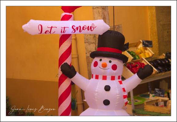 A cheerful inflatable snowman fills the frame, all bright smiles and holiday cheer. He wears a black top hat with a red band, a red-and-white striped scarf, a carrot nose and button eyes, and his stick-like arms are raised as if waving. Beside him a candy-cane-striped pole holds a small sign that reads "Let it snow." The scene is lit with warm, golden light against a plastered wall, and to the right you can glimpse a market stall with crates of produce, which lends the display a cozy, unexpected street-market charm. The whole composition feels playful and festive.