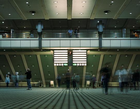 An indoor shot of the International Congress Centre Berlin interior, featuring a prominent green-and-white wall with a large departure information display, illuminated with the number “7” and flight data. Multiple people blurred by motion, walking on the patterned floor of the station. A raised mezzanine features two people observing the scene.
