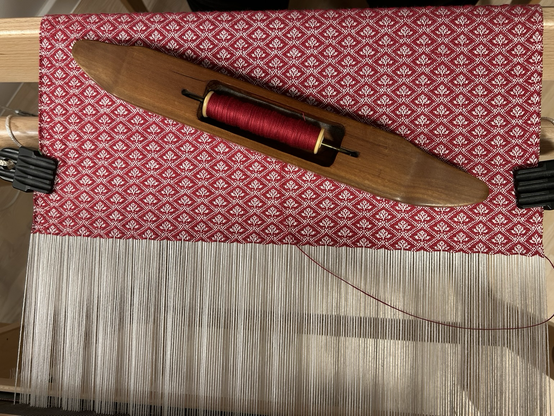 Looking down on handweaving in progress on a hand loom - a complex red twill on a white warp, with a wooden boat shuttle sitting obliquely on the woven fabric