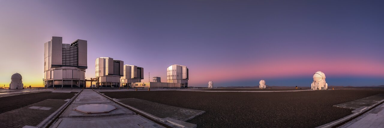 The Paranal Observatory glowing in the light of the setting Sun on the left and a pinkish band on the sky right on the other side. Right below the pink hue, the Earth shadow is cast onto the sky