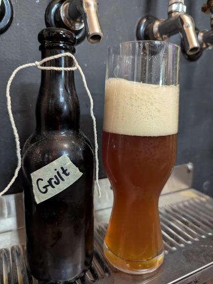 A glass of copper colored beer with a thick head of foam on top stands next to a brown bottle with a masking tape label with the word 'Gruit' written on it. String that formerly held the cork in the bottle hangs down from around the bottle's neck